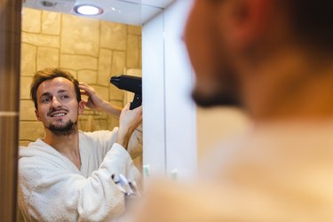 Smiling young receding hairline man in a bathrobe blow drying his hair after taking a shower while looking at himself in the bathroom mirror and touching hair to prevent bad hairlines