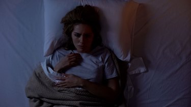 a person with long brown hair wearing a white t-shirt and lying awake in bed because they feel a presence in the room while sleeping