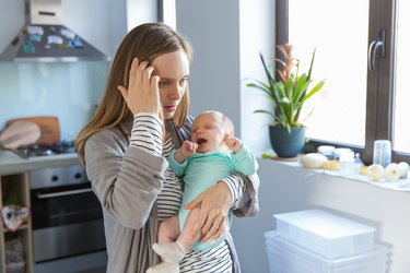 a person with long blonde hair wearing a grey sweater and holding a newborn baby and looking tired and concerned because of postpartum thyroiditis symptoms