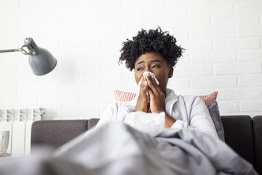 Person with black curly hair lying in bed and blowing their nose because they have the flu with dizziness